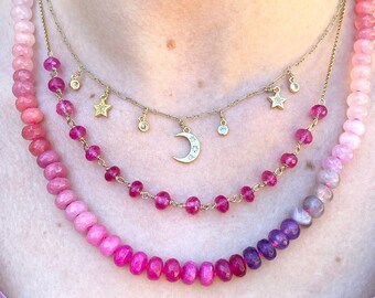 May include: A layered necklace with a crescent moon pendant and star charms. The necklace is made of pink and purple beads.