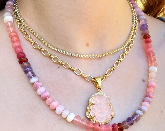 May include: A layered necklace with a pink and purple beaded strand, a gold chain with a pink crystal Buddha pendant, and a gold chain with clear crystals.