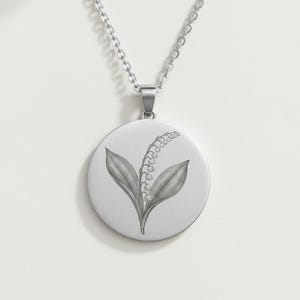 Peut inclure: Un collier argenté avec un pendentif rond gravé d'un motif de muguet. L'illustration de la plante comprend des feuilles et de délicates fleurs en forme de cloche. La chaîne est de style classique, et le pendentif est centré.