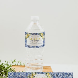 May include: Clear plastic water bottle with a decorative label. The label features the text "She Found Her Main Squeeze" with a lemon and floral design. The label has a blue and white tile pattern. The bottle is on a wooden surface.