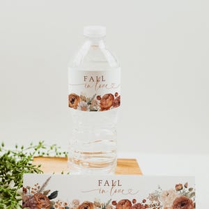 May include: A clear water bottle with a label that reads "Fall in love" in a script font. The label features floral illustrations in shades of orange, brown, and white. A matching floral banner is below the bottle.