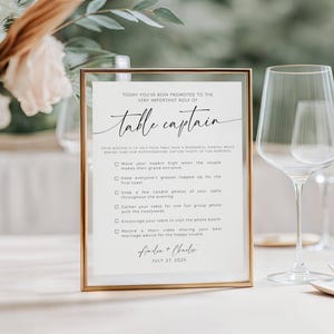 Puede incluir: Un letrero rectangular con marco dorado sobre una mesa con papel blanco que presenta texto negro: TODAY YOU'VE BEEN PROMOTED TO THE VERY IMPORTANT ROLE OF Table captain. Enumera tareas para los invitados e incluye los nombres Amelia + Charles y JULY 27, 202X.