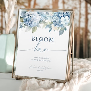 May include: A framed sign with a floral design and the words "BLOOM bar." The sign is in a gold frame and features blue and white flowers at the top. Text on the sign reads "Make a bouquet to take on your way."