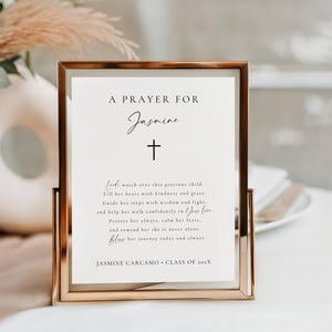 Puede incluir: Una impresión enmarcada con el texto "A Prayer For Jasmine" y un símbolo de cruz. La impresión incluye una oración y el nombre "Jasmine Carcamo" con "Class of 202X". El marco es de color oro rosa metálico.
