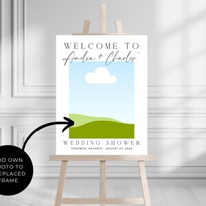 May include: A wedding shower sign on a wooden easel. The sign has a blue sky and green landscape graphic. Text reads "WELCOME TO Amelia + Charles" and "WEDDING SHOWER TORONTO, ONTARIO - AUGUST 27, 202X". A black circle with an arrow points to the sign.