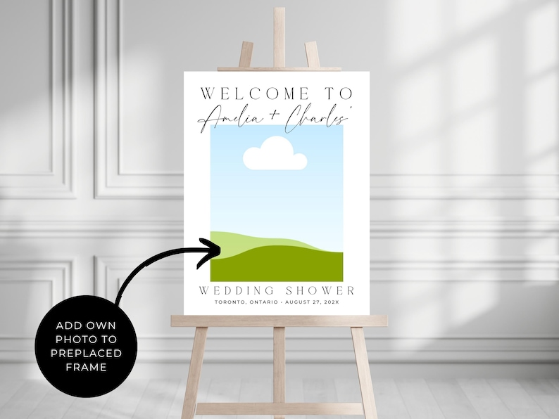 May include: A wedding shower sign on a wooden easel. The sign has a blue sky and green landscape graphic. Text reads "WELCOME TO Amelia + Charles" and "WEDDING SHOWER TORONTO, ONTARIO - AUGUST 27, 202X". A black circle with an arrow points to the sign.