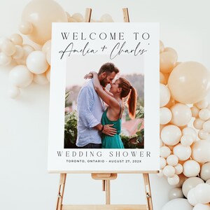May include: A white wedding shower sign on a wooden easel. The sign reads "Welcome to Amelia + Charles" above a photo of a couple embracing. The words "Wedding Shower" and event details are below the photo. Balloons decorate the background.
