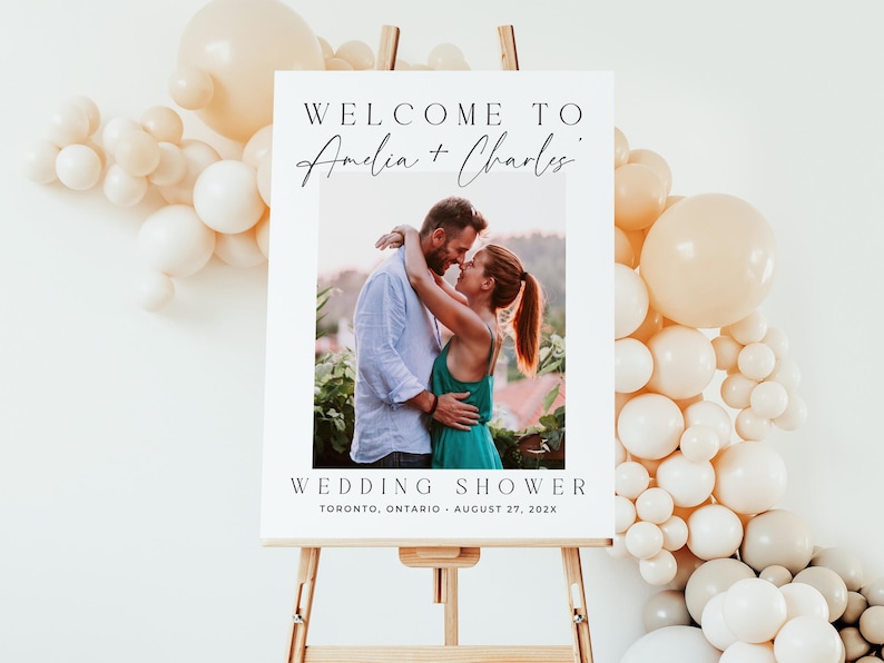 May include: A white wedding shower sign on a wooden easel. The sign reads "Welcome to Amelia + Charles" above a photo of a couple embracing. The words "Wedding Shower" and event details are below the photo. Balloons decorate the background.