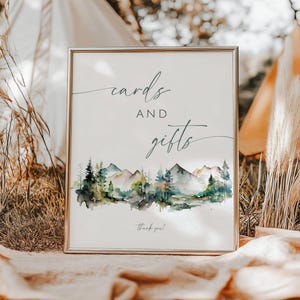 May include: A rectangular sign in a thin gold frame featuring the text cards AND gifts in a mix of serif and script fonts. Below the text is a watercolor illustration of a mountain range with green pine trees and a small thank you at the bottom.