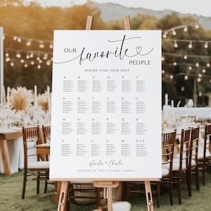May include: A white seating chart on an easel with the text "Our Favorite People" and a list of names. The sign includes the names Amelia and Charlie, and the date November 16, 2025, in Calgary, Alberta. The background shows a wedding reception.