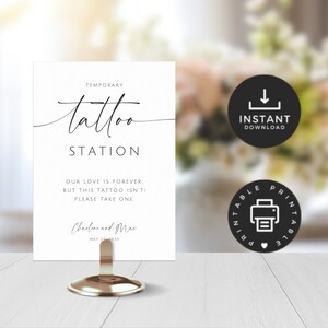 May include: A white sign for a temporary tattoo station, featuring elegant script lettering. The sign says "Temporary Tattoo Station" and includes the text "Our love is forever, but this tattoo isn't, please take one." The sign is held by a gold metal stand.