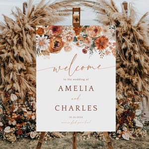 May include: A wedding welcome sign with floral arrangements in shades of orange, cream, and burgundy. The sign reads "welcome" and the names "AMELIA and CHARLES" with the date "10.25.2023". The sign is on an easel.