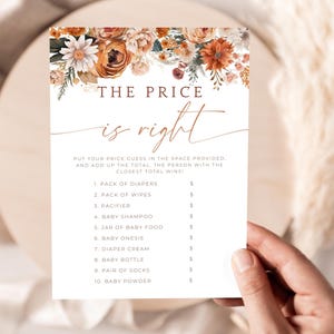 May include: A baby shower game card titled "The Price is Right" with a floral design in shades of orange, brown, and white. The card lists baby items like diapers and baby food, with spaces for price guesses. The card is held by a person.
