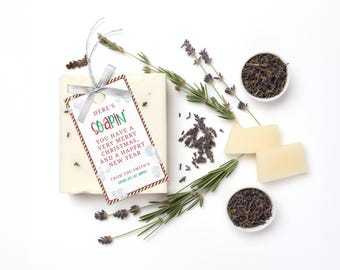 Op de afbeelding: Een witte zeep met een rood, groen en wit cadeau-etiket waarop staat "Here's Soapin' You have a very merry Christmas, and a happy new year from the Smith's". De zeep is omringd door lavendeltakjes en gedroogde lavendel.
