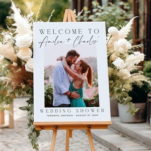 May include: A white wedding welcome sign on a wooden easel. The sign features the text "WELCOME TO Amelia + Charles" and a photo of a couple embracing. Below the photo, it reads "WEDDING SHOWER" and the event details.