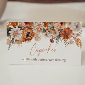 Op de afbeelding: Een rechthoekig bord met een bloemenontwerp en het woord "Cupcakes" in een scriptlettertype. Het bord bevat ook de tekst "vanille met botercrème glazuur". Het bloemstuk bevat oranje, witte en rode bloemen.