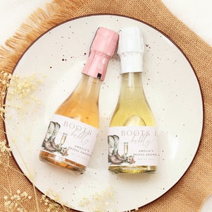 Puede incluir: Dos mini botellas de champán con etiquetas rosas y blancas. Las etiquetas presentan una ilustración de acuarela de botas de vaquero, una copa de champán y detalles florales. El texto en las etiquetas dice "Boots & bubbly" y "Amelia's Bridal Shower".