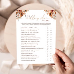 May include: A wedding shoe game card with floral accents in shades of orange and white. The card features a list of questions for the couple to answer. The text "The Wedding Shoe Game" is at the top.