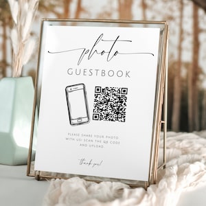 May include: White sign with black text and graphics. The sign reads "photo guestbook". A black and white illustration of a smartphone and a QR code are shown. The text below reads "Please share your photo with us! Scan the QR code and upload. Thank you!"