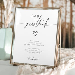 May include: A framed sign with the words "BABY guestbook" in elegant script, a heart symbol, and instructions to sign. The sign is in a gold frame, with a soft, neutral background.