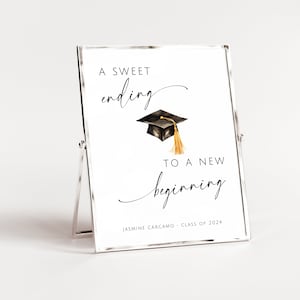 May include: A white sign with black text that reads "A sweet ending to a new beginning". A black graduation cap with a gold tassel is above the text. The sign also includes the text "Jasmine Carcamo - Class of 2024".
