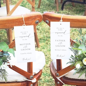May include: Two white signs with black text hang from the back of two wooden chairs. The signs say "This seat is kindly reserved for the Mother of the Groom, Camilla Adams" and "This seat is kindly reserved for the Father of the Groom, Michael Adams". Both signs have a small floral arrangement behind them.