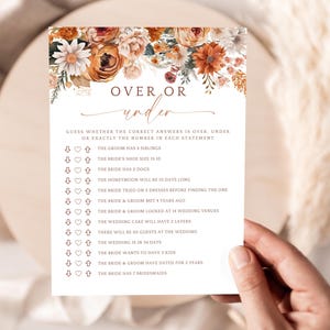May include: A wedding game card with floral accents in orange and white. The text "OVER OR under" is at the top. The card includes questions about the bride and groom, such as the number of siblings and bridesmaids. The card is held by a person.