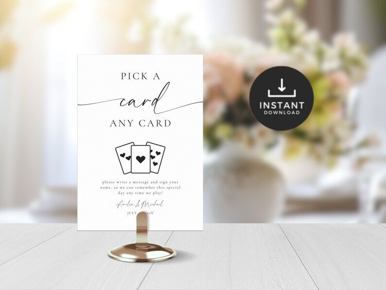 Op de afbeelding: Een witte kaart met zwarte tekst die "PICK A card ANY CARD" zegt. Er zijn drie zwarte speelkaarten met harten erop afgebeeld. De tekst onder de kaarten luidt "Schrijf een bericht en teken je naam, zodat we deze speciale dag kunnen herinneren wanneer we spelen!" gevolgd door de namen "Finder & Mubad" en de datum "JULI".