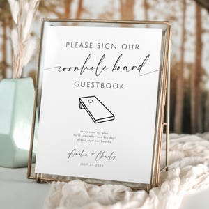 May include: A framed sign with the text "PLEASE SIGN OUR cornhole board GUESTBOOK". The sign includes a cornhole board illustration and the names "Amelia + Charles" with a date. The frame is gold.