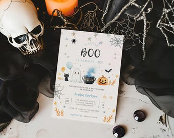 May include: A white card with a Halloween baby shower invitation. The card features a ghost, a black cat, a cauldron, and a pumpkin. The text on the card reads "A Little Boo is Almost Due!" and includes the date, time, and location of the shower.