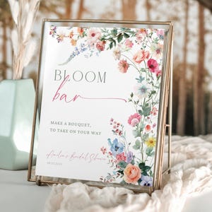 Puede incluir: Un letrero enmarcado con las palabras "Bloom Bar" e ilustraciones florales. El letrero incluye el texto "Make a bouquet, to take on your way" y "Amelia's Bridal Shower 08.20.2024". El marco es de color dorado.