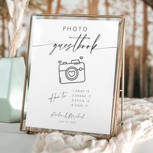 以下が含まれることがあります： カメラの中にハートが描かれたシンプルな線画と、黒い文字が書かれた白い看板。看板には「PHOTO guestbook」と「How to: 1. SNAP IT 2. SHAKE IT 3. STICK IT 4. SIGN IT」と「Anden & Mubad JULY 27 202X」と書かれている。