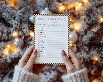 May include: A Christmas Song emoji pictionary card held by a person. The card has a white background with red and green striped borders. The text "Christmas Song" is at the top, with "emoji pictionary" below. The card features a list of emojis.