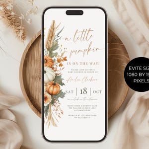 May include: A smartphone displaying a digital baby shower invitation with the text A little pumpkin IS ON THE WAY! The design features watercolor illustrations of orange and white pumpkins, dried grasses, and autumn leaves on a white background.