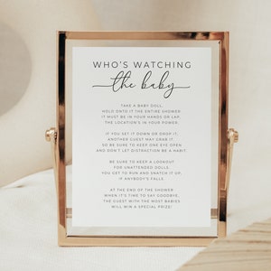 May include: A white framed sign with gold trim, with the text "WHO'S WATCHING the baby" and instructions for a baby shower game. Guests must hold a baby doll for the duration of the shower, and the guest with the most dolls at the end wins a prize.