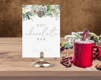 Könnte beinhalten: Ein weißes Schild mit grünen und weißen floralen Akzenten lautet "HOT chocolate BAR warm up here!"  Das Schild steht auf einem goldenen Ständer und eine rote Tasse mit Marshmallows und einem Lebkuchenmann steht im Vordergrund.