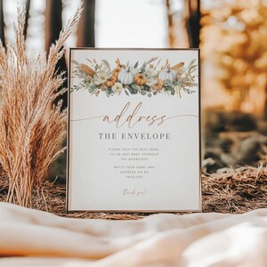 May include: A rectangular sign with a thin gold frame featuring a watercolor illustration of white and orange pumpkins, greenery, and dried wheat at the top. The text reads: address THE ENVELOPE, PLEASE HELP THE BUSY MOM-TO-BE AND MAKE YOURSELF THE ADDRESSEE, WRITE YOUR NAME AND ADDRESS ON AN ENVELOPE, Thank you!