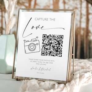 Op de afbeelding: Een wit bord met zwarte tekst en een zwart-witte QR-code. De tekst luidt "Capture the Love" in een gestileerd cursief schrift. Onder de QR-code staat de tekst "Deel uw foto's met ons, scan gewoon de QR-code en upload ze" gevolgd door de namen "Andia & Mubad" en de datum "27 juli 2024".