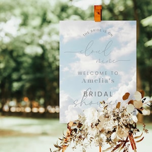 May include: A sign with the text "THE BRIDE IS ON cloud nine WELCOME TO Amelia's BRIDAL shower" against a sky background. The sign is decorated with white and brown flowers and greenery. The sign is held up by a wooden stand.