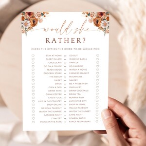 Puede incluir: Una tarjeta de juego para la despedida de soltera con el texto "Would She Rather?" en una fuente decorativa. La tarjeta presenta una lista de opciones para que la futura novia elija, con acentos florales en las esquinas. La tarjeta está en manos de una persona.