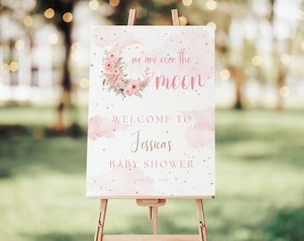 May include: A pink and white baby shower welcome sign with a crescent moon and floral design. The text reads "We are over the moon. Welcome to Jessica's Baby Shower. March 21, 2023."