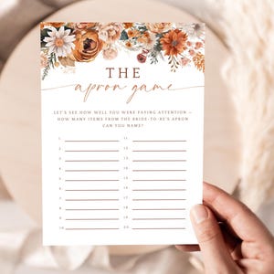 May include: A white card with the text "THE apron game" and floral accents. The card is designed for a bridal shower game, with prompts to list items from the bride's apron. The card is held by a person.
