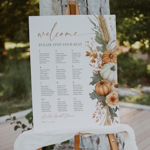 May include: A welcome sign for a bridal shower, displayed on an easel. The sign features a floral design with pumpkins and fall foliage. The text reads "Welcome" and "Please Find Your Seat", with a list of names below.