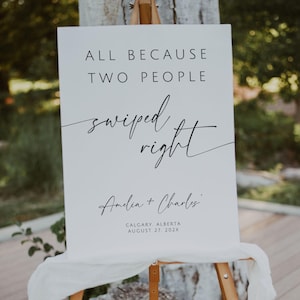 May include: A white sign on a wooden easel with the text "ALL BECAUSE TWO PEOPLE swiped right." Below the text are the names "Amelia + Charles" and the date and location of the event.