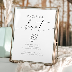 May include: A framed sign with the words "PACIFIER hunt" in elegant script. The sign includes text about a game where pacifiers are hidden, and the person who finds the most wins. The frame is gold-colored, and the background is blurred.