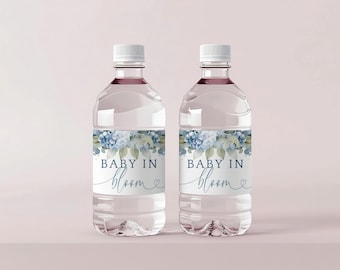 May include: Two clear plastic water bottles with white caps, each featuring a label with a watercolor illustration of blue hydrangea flowers and green leaves, with the text "BABY IN bloom" printed in dark blue and script lettering.