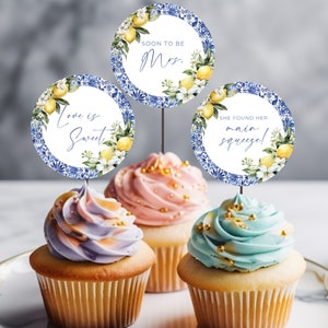 May include: Three cupcakes with pastel frosting and decorative toppers. The toppers are round with a blue and white floral design, lemons, and text that reads "Love is Sweet", "Soon to be Mrs.", and "She found her main squeeze!"