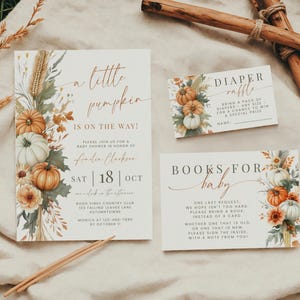 May include: A baby shower invitation set featuring a large invitation, a diaper raffle card, and a books for baby card. The stationery has a cream background with watercolor illustrations of pumpkins, wheat, and autumn leaves in orange and green.