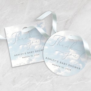 May include: Two baby shower thank you tags with a sky-blue cloud design. One tag is square, the other is round. Both have the text "Thank you!" and "ASHLEY'S BABY SHOWER MARCH 16, 202X" in a delicate script font. A white ribbon is attached.