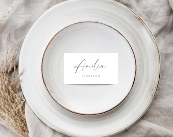 Può includere: Un segnaposto bianco con calligrafia nera che recita "Amelia Clarkson". Il segnaposto è su un piatto bianco.